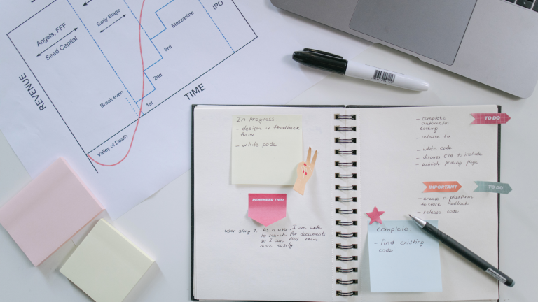 An open notebook sits on a desk with part of a laptop, sticky notes, and other paperwork visible.
